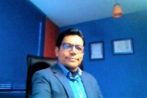 foto con imagen del rostro de Luis Venegas Chalen, sonriente, en traje formal, en su consultorio oficina clínica de hipnosis, en la pared se muestra sus diplomas y título que lo autorizan como hipnoterapeuta profesional.